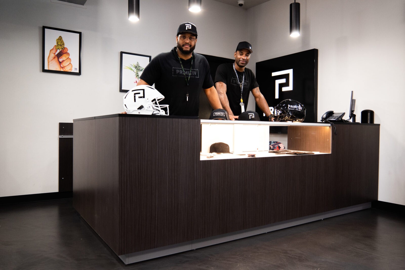 Inside a sleek Primitiv shop, two men in branded attire stand behind a dark counter displaying a white football helmet and other merchandise, with framed artwork on the wall.