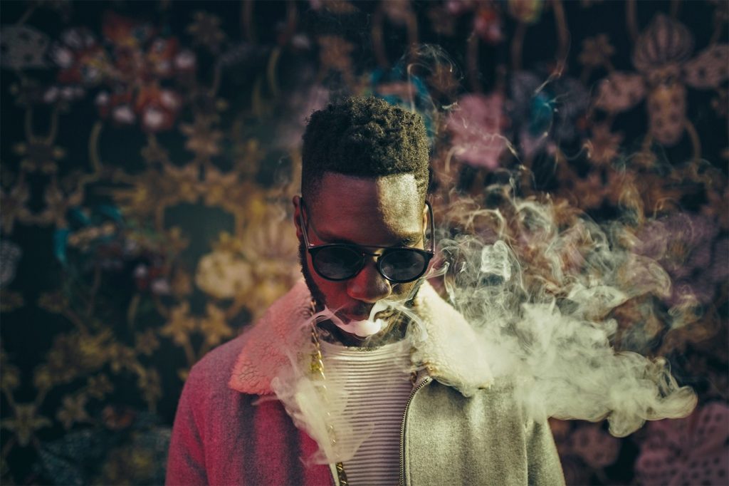 Stylized portrait of a man wearing sunglasses and a pink jacket, exhaling smoke in front of a dark floral-patterned background.