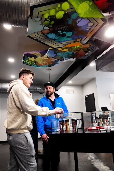A customer and a budtender engaging in conversation at a Primitiv dispensary counter, showcasing a sleek interior design with colorful modern art displayed on the ceiling.