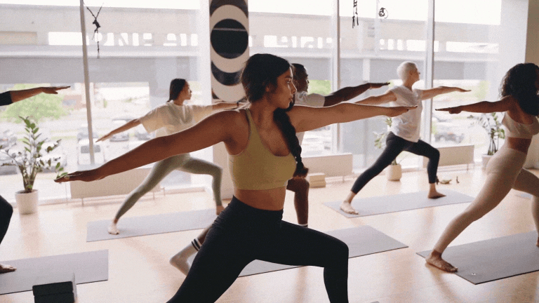 Animated GIF showing a group of people doing yoga in a bright studio; a woman in a yellow sports bra leads the group in a warrior pose while others follow on mats.