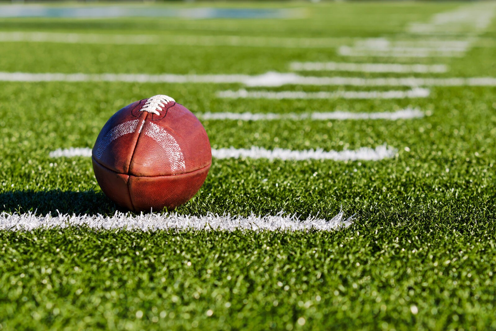 A close-up view of a football resting on the field, with the white yard lines clearly visible on the green turf.